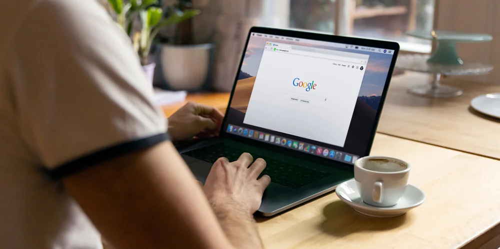 Pessoa utilizando um notebook com a página inicial do Google aberta, em um ambiente de trabalho com café ao lado, representando pesquisa e estratégias digitais.